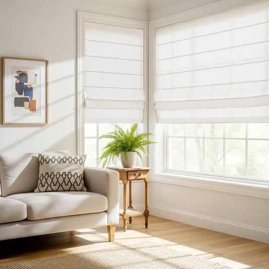 Motorized Roman shades in bright living room corner with beige sofa, patterned cushion, small wooden side table with potted plant, and large windows overlooking greenery