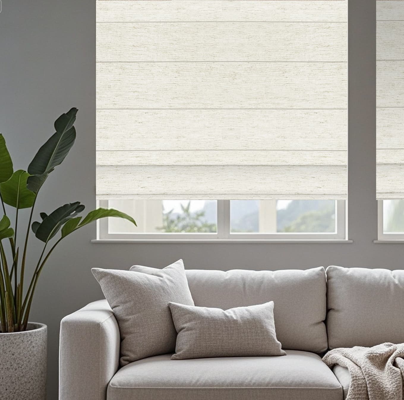 Motorized Roman shade in modern living room with light gray sofa, soft cushions, indoor plant, linen and window overlooking greenery